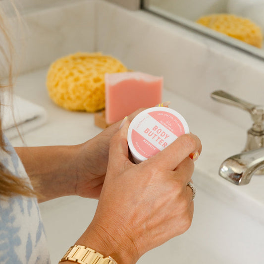 Model opening travel size body butter in bathroom with bar soap on soap dish in background