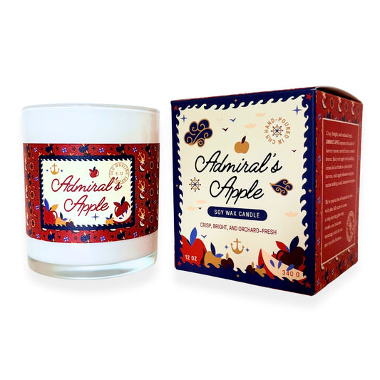 A soy wax candle in a clear glass jar with a white wax fill and a decorative red, navy, and cream label reading 'Admiral's Apple,' shown next to its matching product box on a white background.