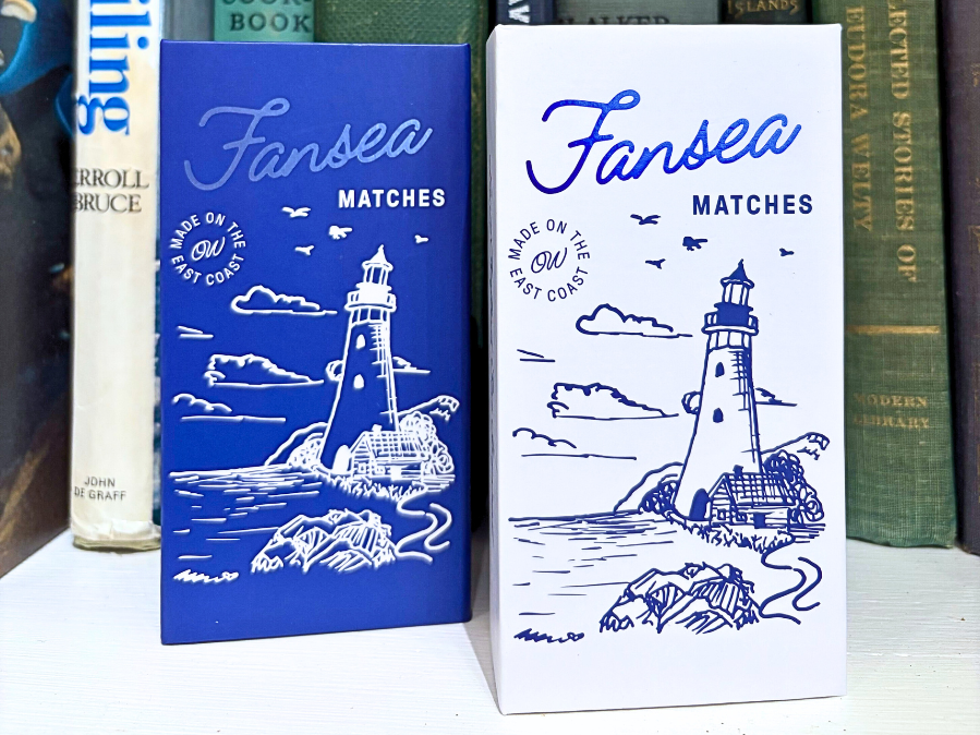Two matchboxes with a lighthouse design on a bookshelf.