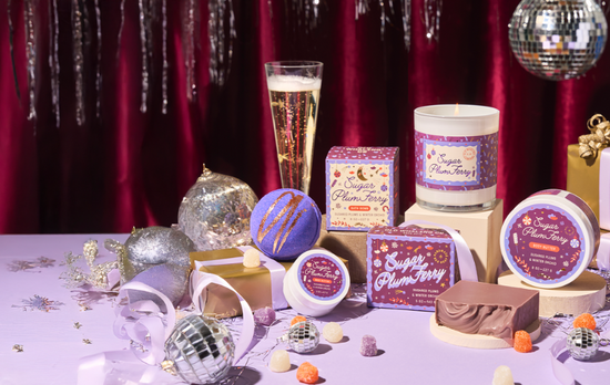 Table with various holiday products including candles, soaps, and bath products against a festive background with disco balls