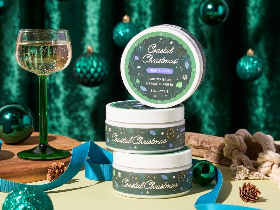 Three jars of 'Coastal Christmas' body butter stacked on a table with a glass of wine and festive decorations in the background.