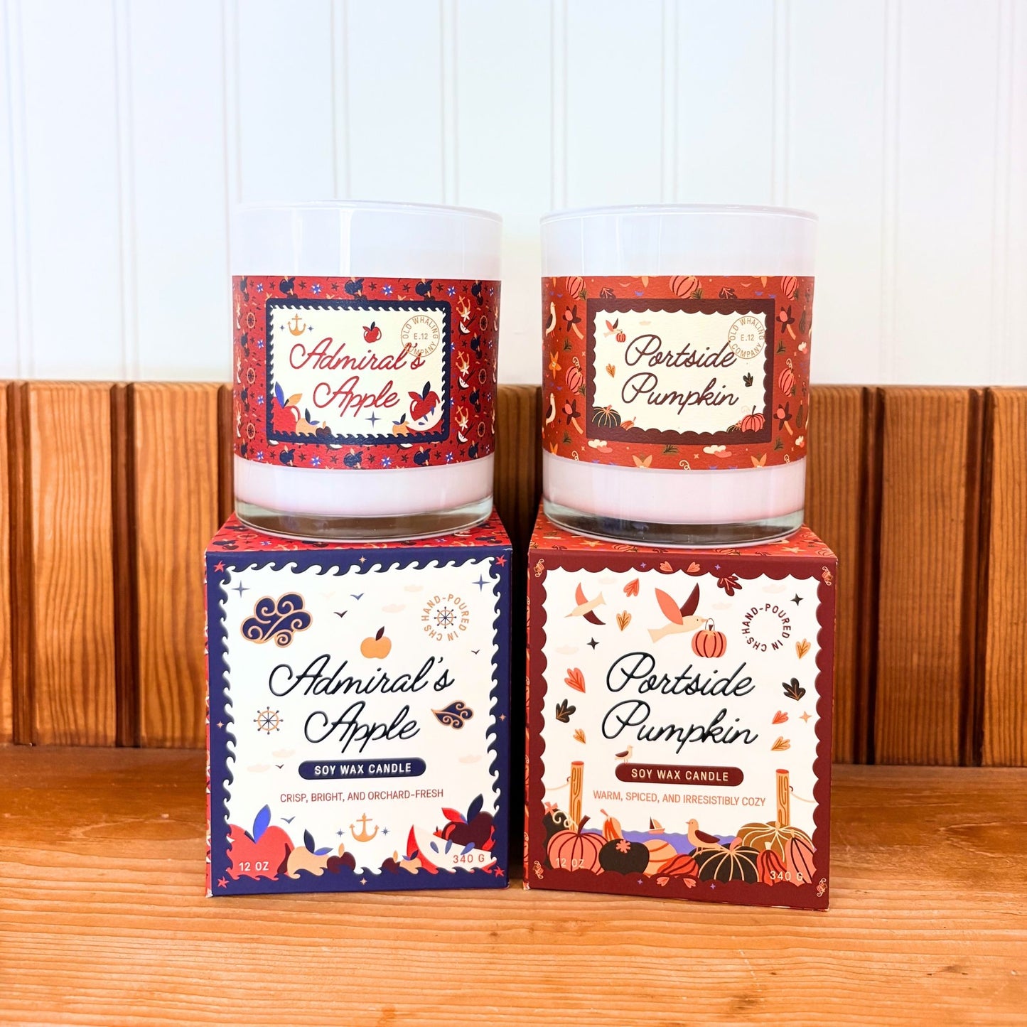 Two scented soy wax candles in clear glass jars with autumn-themed labels, displayed on top of their matching illustrated boxes on a wooden surface against a white paneled wall.