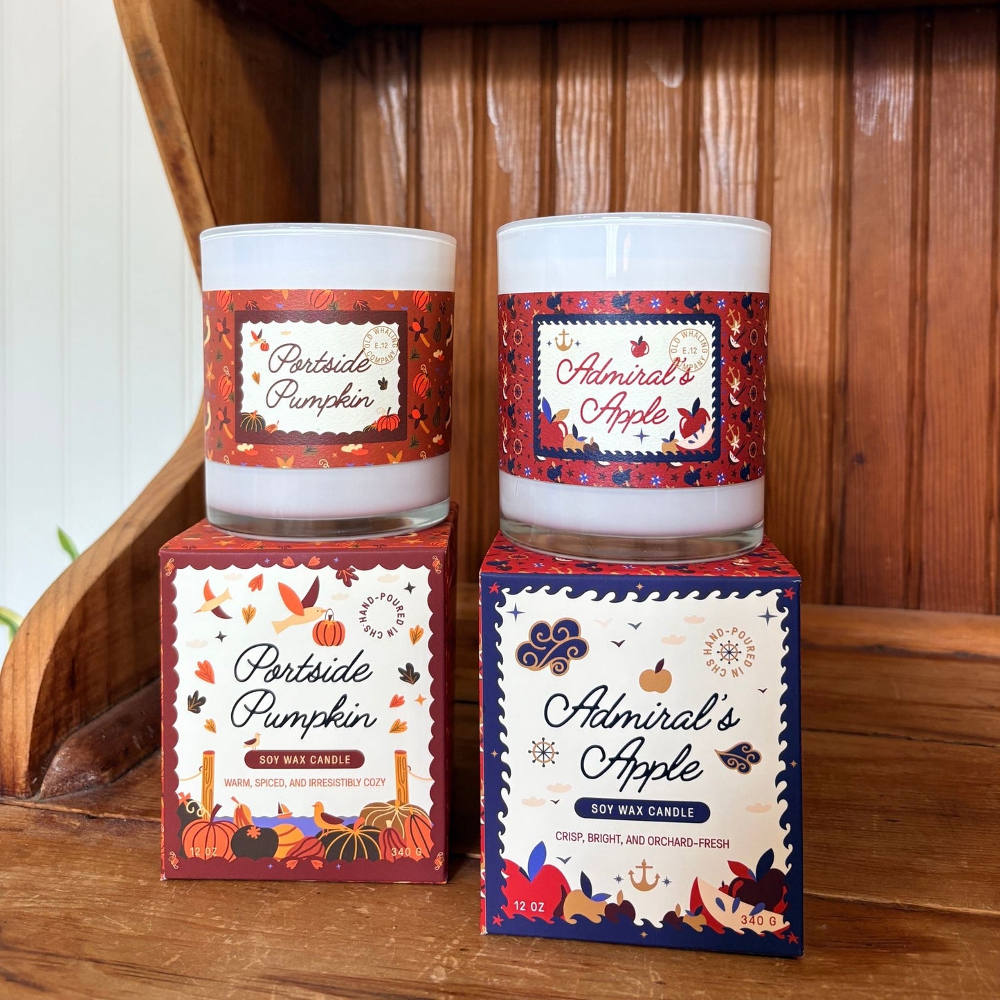 Two scented soy wax candles in glass jars with red and orange autumn-themed labels, displayed on top of their matching retail boxes on a wooden surface.