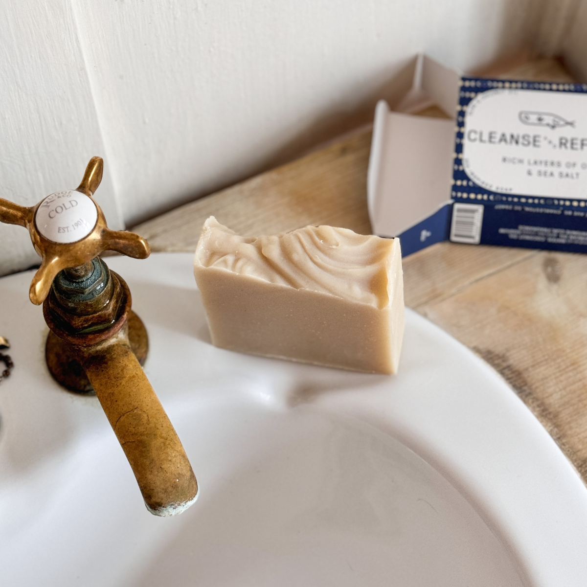 A bar of creamy beige soap sits on a white ceramic sink next to a brass faucet, with its blue and white packaging box visible in the background on a wooden surface.