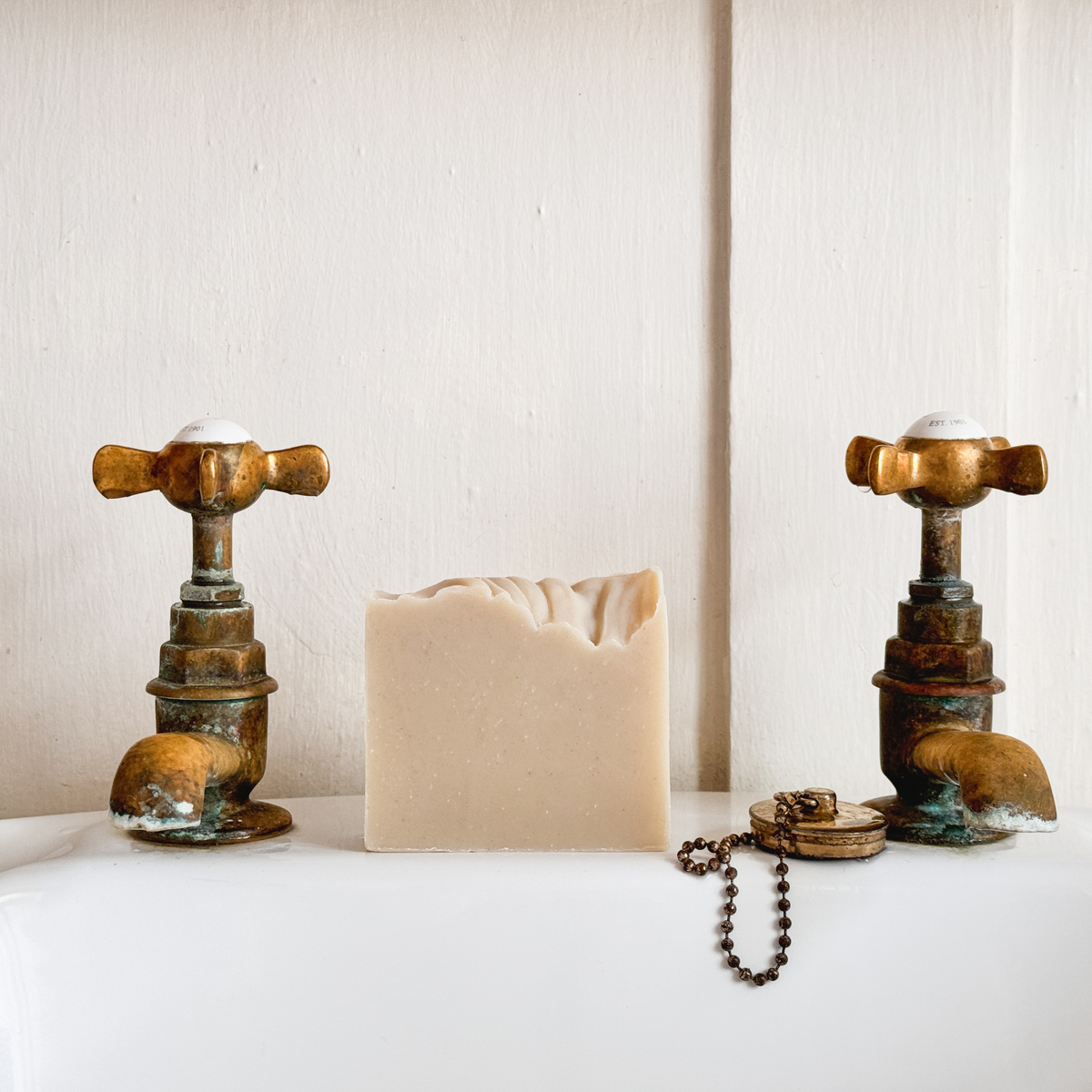 A rectangular bar of creamy beige soap stands centered on a white surface between two antique-style brass faucet fixtures, with a small chain accessory in front. The background is a plain off-white wall.