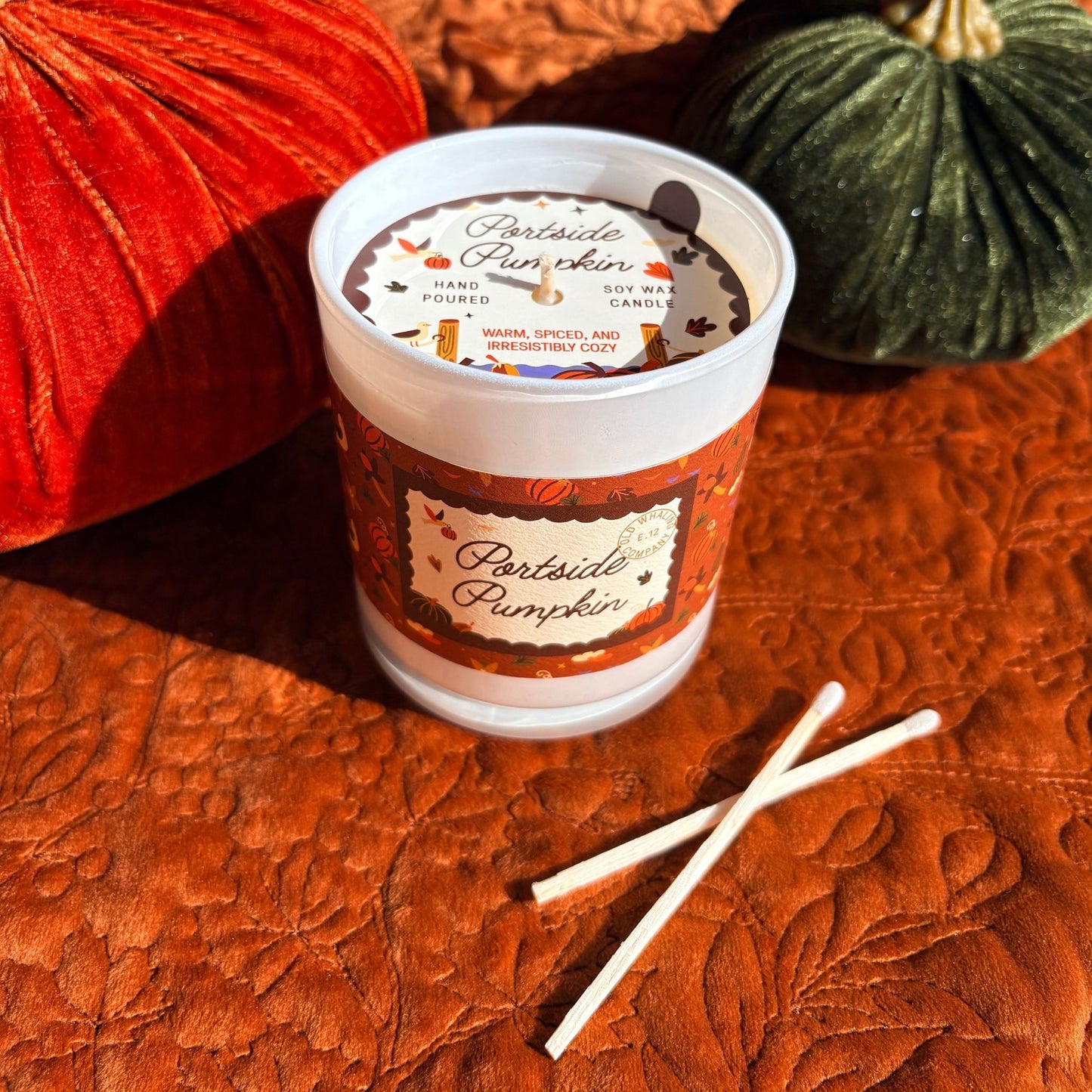 A hand-poured soy wax candle in a jar with a white dust cover and a fall-themed label reading 'Portside Pumpkin.' The candle sits on a textured rust-orange surface with decorative pumpkins in the background and two matchsticks placed in front.