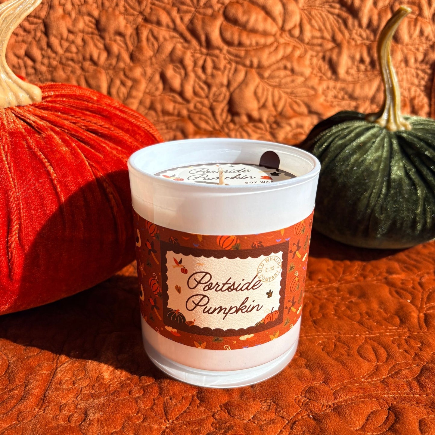 A frosted glass jar candle labeled 'Portside Pumpkin' sits on an orange quilted blanket, styled with a red velvet pumpkin on the left and a green velvet pumpkin on the right.