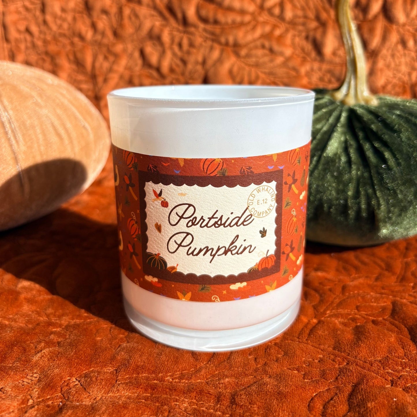 A frosted glass jar candle labeled 'Partside Pumpkin' sits on a textured rust-orange surface with decorative pumpkins in the background.