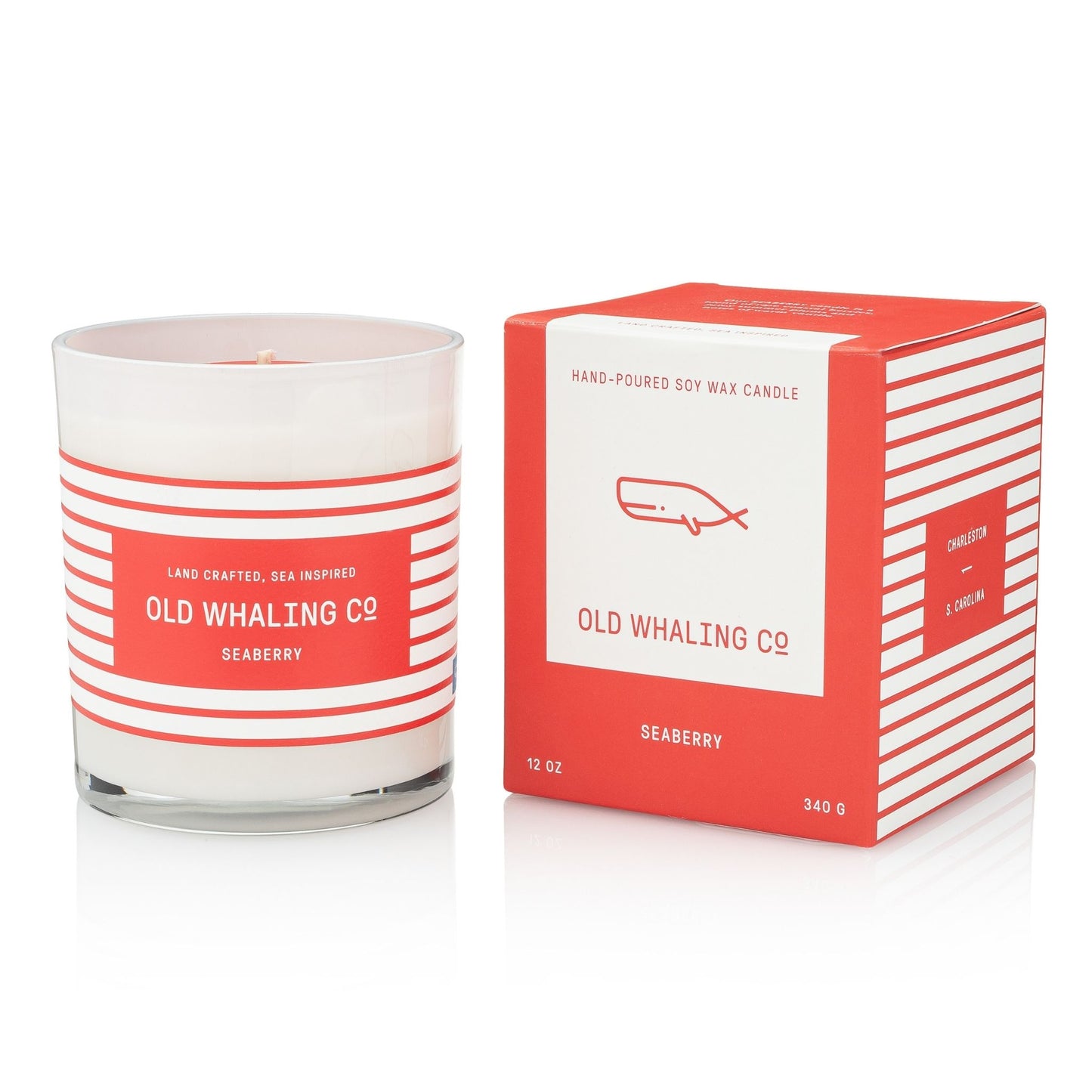 A white soy wax candle with a red and white striped label sitting next to it's matching box from brand Old Whaling Company and scent name Seaberry