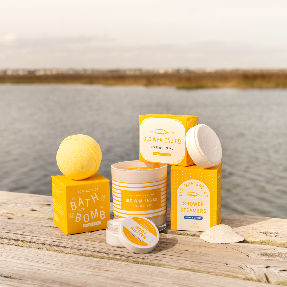 A curated collection of Old Whaling Co. bath and body products arranged on a weathered wooden dock with a calm body of water and distant shoreline in the background.