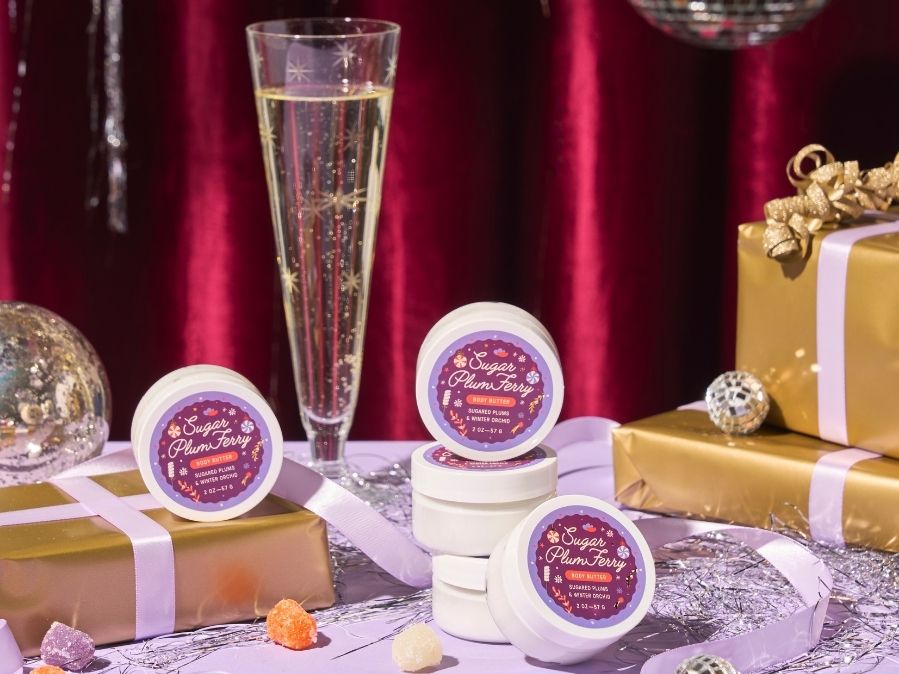 Jars of 'Sugar Plum Fairy' body butter with a glass of champagne and wrapped gifts on a festive background.