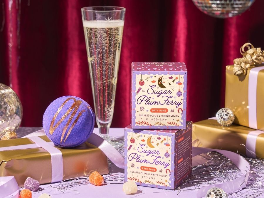 Two boxes of 'Sugar Plum Fairy' candy, a glass of champagne, and decorative items on a table with a red curtain background.