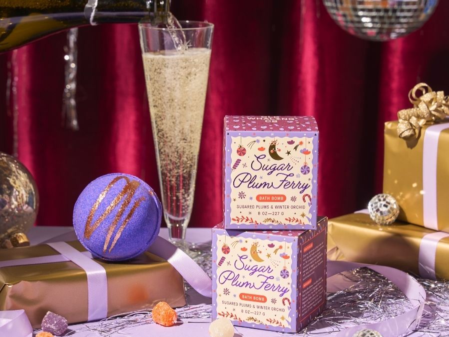 Two 'Sugar Plum Fairy' bath bombs with a glass of champagne and wrapped gifts on a red background.