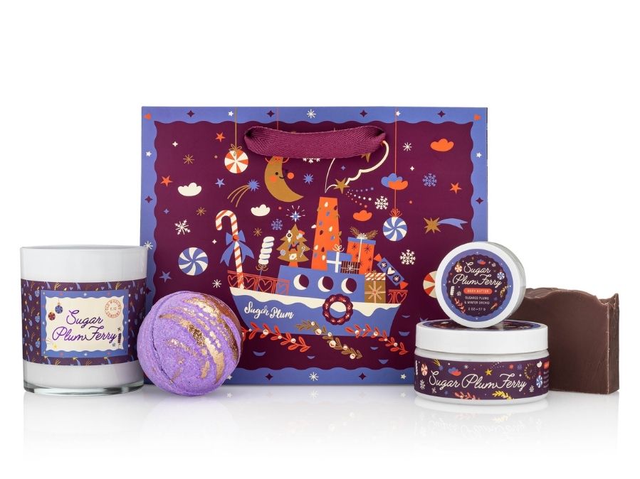 Set of 'Sugar Plum Fairy' products including a candle, bath bomb, and body butter with a decorative gift bag in the background.