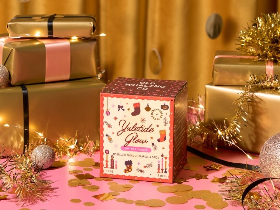 Yuletide Glow candle box surrounded by gold presents and festive decorations on a pink surface.