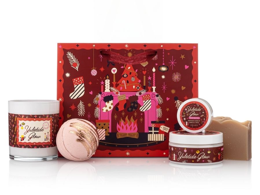 Set of holiday-themed products including a candle, bath bomb, and body butter with a decorative gift bag tote in the background.